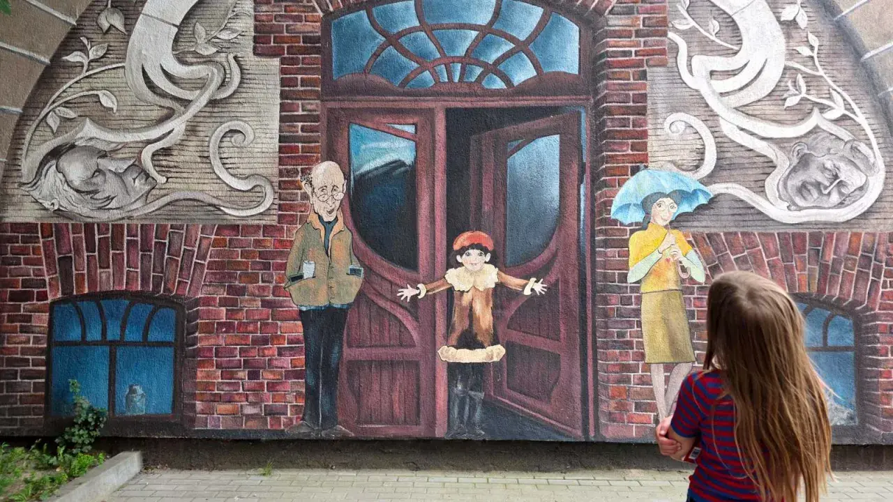 Dziecko patrzy na mural z otwartymi drzwiami, sugerujący jak uciec i zacząć nowe życie. Obok stoją postacie: starszy pan, dziecko i kobieta z parasolką.