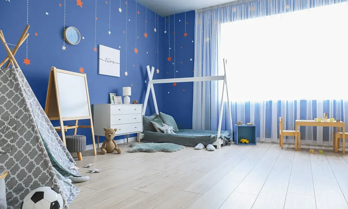 Pokój dziecka z niebieskimi, malowanymi ścianami ozdobionymi gwiazdkami. W rogu stoi namiot tipi, obok łóżko i komoda.