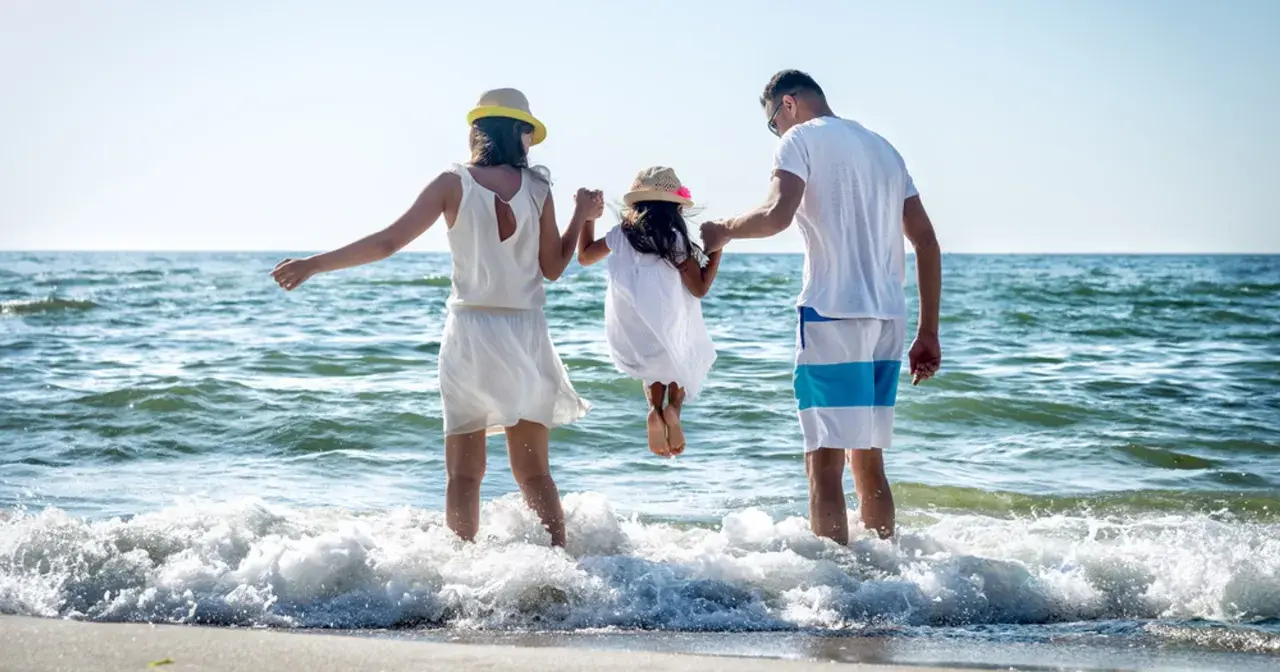 Rodzina na plaży, rodzice trzymają córkę za ręce, która skacze w falach. To kwintesencja bycia szczęśliwym.