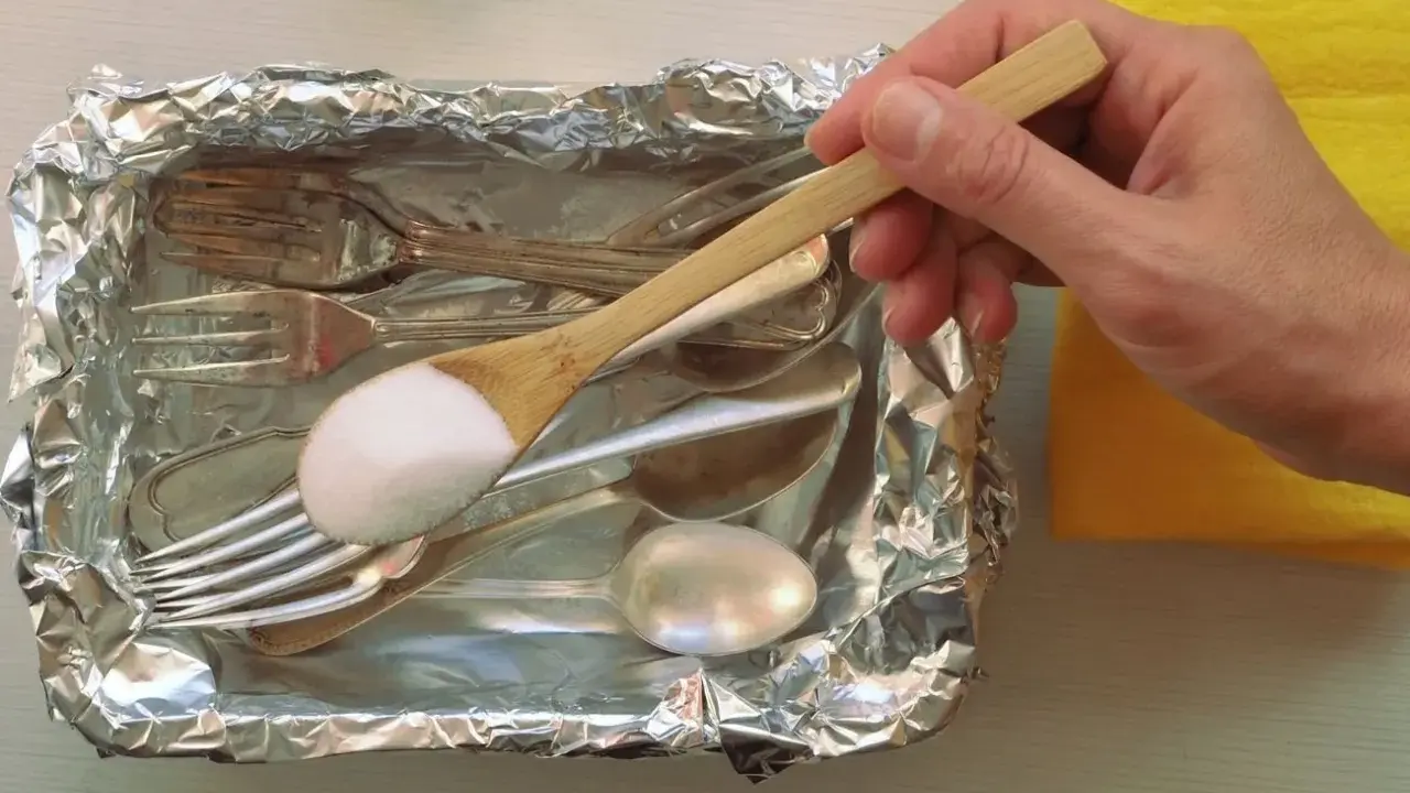 Łyżka z sodą oczyszczoną nad tacą z folii aluminiowej, pełną sztućców do czyszczenia srebra.