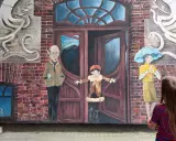 Dziecko patrzy na mural z otwartymi drzwiami, sugerujący jak uciec i zacząć nowe życie. Obok stoją postacie: starszy pan, dziecko i kobieta z parasolką.