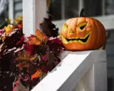 Uśmiechnięta dynia z wyciętymi oczami i zębami, otoczona jesiennymi liśćmi i pajęczyną. Idealne dekoracje z dyni ozdobnej na Halloween.