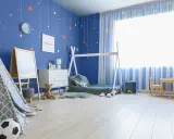Pokój dziecka z niebieskimi, malowanymi ścianami ozdobionymi gwiazdkami. W rogu stoi namiot tipi, obok łóżko i komoda.