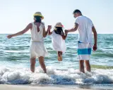 Rodzina na plaży, rodzice trzymają córkę za ręce, która skacze w falach. To kwintesencja bycia szczęśliwym.