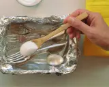 Łyżka z sodą oczyszczoną nad tacą z folii aluminiowej, pełną sztućców do czyszczenia srebra.