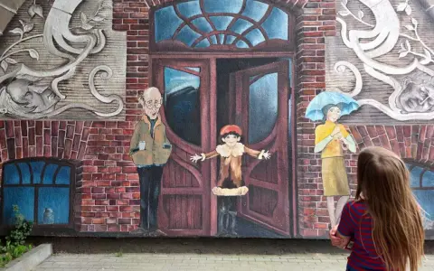Dziecko patrzy na mural z otwartymi drzwiami, sugerujący jak uciec i zacząć nowe życie. Obok stoją postacie: starszy pan, dziecko i kobieta z parasolką.