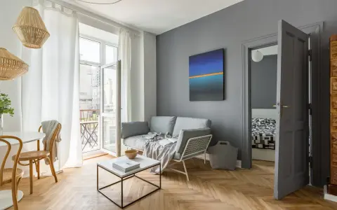 Przytulny, mały salon z szarą kanapą, stolikiem kawowym i obrazem na ścianie. Drewniana podłoga i wiklinowe lampy dodają ciepła.