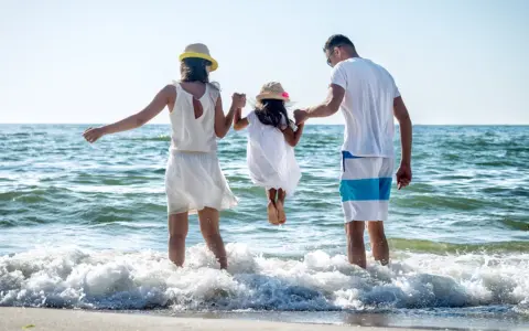 Rodzina na plaży, rodzice trzymają córkę za ręce, która skacze w falach. To kwintesencja bycia szczęśliwym.