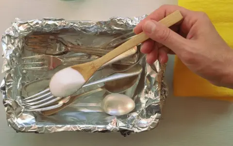 Łyżka z sodą oczyszczoną nad tacą z folii aluminiowej, pełną sztućców do czyszczenia srebra.