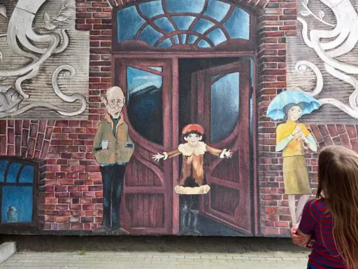 Dziecko patrzy na mural z otwartymi drzwiami, sugerujący jak uciec i zacząć nowe życie. Obok stoją postacie: starszy pan, dziecko i kobieta z parasolką.