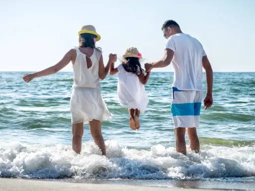Rodzina na plaży, rodzice trzymają córkę za ręce, która skacze w falach. To kwintesencja bycia szczęśliwym.