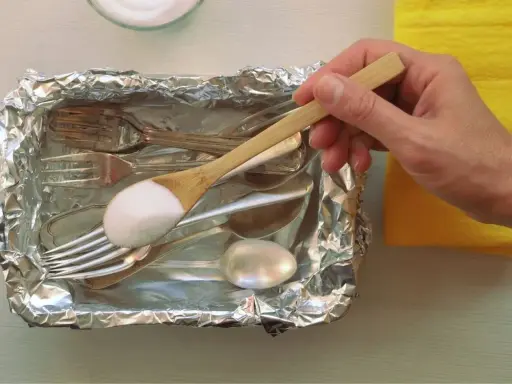 Łyżka z sodą oczyszczoną nad tacą z folii aluminiowej, pełną sztućców do czyszczenia srebra.