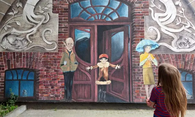 Dziecko patrzy na mural z otwartymi drzwiami, sugerujący jak uciec i zacząć nowe życie. Obok stoją postacie: starszy pan, dziecko i kobieta z parasolką.