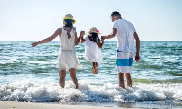 Rodzina na plaży, rodzice trzymają córkę za ręce, która skacze w falach. To kwintesencja bycia szczęśliwym.