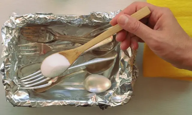 Łyżka z sodą oczyszczoną nad tacą z folii aluminiowej, pełną sztućców do czyszczenia srebra.