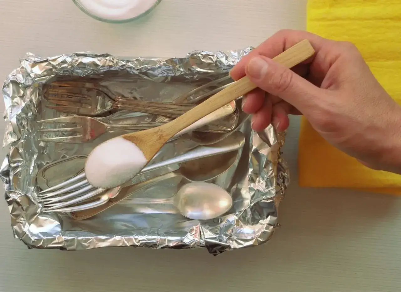 Łyżka z sodą oczyszczoną nad tacą z folii aluminiowej, pełną sztućców do czyszczenia srebra.